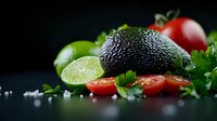Fresh ripe avocado with sliced lime, cherry tomatoes and herbs on dark background with sea salt crystals, dramatic food photography composition.