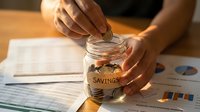 Hands placing coins into glass jar labeled savings with financial documents and charts on wooden desk for money management and budgeting planning.