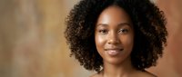 Young African American woman with natural curly hair smiling warmly at camera in soft indoor lighting for professional headshot and portrait photography.