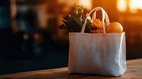 Fresh groceries in white eco-friendly shopping bag with fruits, vegetables and greens on wooden table against warm sunset light through window.