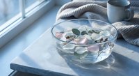 Glass bowl with floating rose petals and eucalyptus leaves on marble surface near window with soft natural light creating peaceful spa atmosphere.