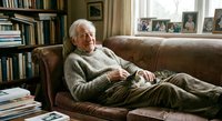 Elderly Caucasian man relaxing on leather sofa in cozy home library with bookshelves and family photos, comfortable retirement lifestyle concept.