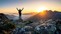 Silhouette of person standing on rocky mountain peak with arms raised at sunrise, celebrating achievement and freedom with golden sunlight and misty valleys below.