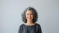Mature woman with gray curly hair and warm smile wearing dark casual shirt against neutral background for portrait use.