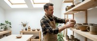 Middle-aged male artisan with salt-and-pepper hair and light skin tone, wearing a brown apron over a checkered shirt, carefully examining a handmade ceramic bowl in a bright, minimalist pottery studio with wooden shelves and plants.
