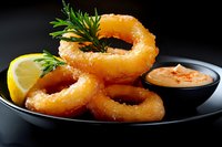 Crispy golden onion rings served on black plate with spicy dipping sauce, fresh lemon slice and parsley garnish against dark background. Restaurant appetizer.
