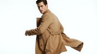 Young Caucasian man wearing tan trench coat, looking over shoulder against white background. Suitable for fashion, lifestyle, and professional business concepts.