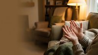 Person relaxing at home with hands raised in cozy living room with warm lamp lighting and comfortable furniture for wellness and self-care concepts.