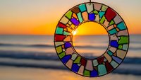 Colorful stained glass mosaic circle suncatcher hanging against golden sunset ocean backdrop creating rainbow light reflections for home decor projects.
