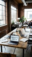 Innovative startup coworking space with laptops on rustic wooden table, brick walls, and colleagues collaborating in background.