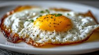 Close-up of a sunny side up fried egg with crispy edges on white plate, sprinkled with black pepper and fresh herbs for breakfast or brunch.