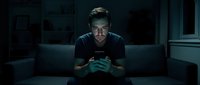 Young man with dark hair and light skin tone, wearing black t-shirt, sitting on couch in dimly lit room holding gaming controller. Gamer focused on video game competition.