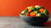 Fresh mixed vegetables including broccoli florets and cherry tomatoes in ceramic bowl on wooden surface against vibrant orange background for healthy cooking.