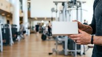 Hands holding blank white card in modern gym facility with exercise equipment and blurred background, suitable for fitness promotions and wellness marketing.