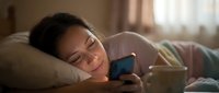 Young woman of East Asian descent relaxing on bed with smartphone, warm natural lighting streaming through window, casual leisure and mobile technology use.