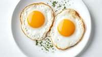 Two sunny side up eggs with crispy edges and bright yellow yolks garnished with fresh herbs served on white plate, view from above on light background.