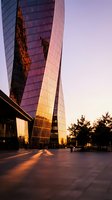 Modern glass skyscraper facade illuminated by golden sunset light, casting warm reflections on urban plaza pavement during twilight hour for architectural and cityscape projects.