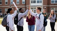 Diverse group of college students celebrating together with high fives on university campus courtyard, showing teamwork and friendship among peers.