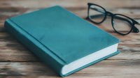 Teal hardcover book with black-rimmed glasses on rustic wooden surface for reading, education, literature, study concepts and academic lifestyle designs.