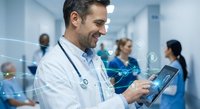 Smiling male doctor using digital tablet with holographic medical interface in modern hospital corridor with healthcare team working in background.