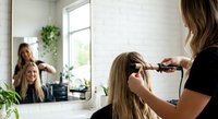 Professional hairstylist curling blonde woman's hair with curling iron in modern bright salon with natural light and plants creating relaxed atmosphere.