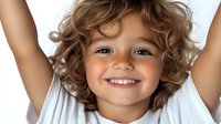 Joyful young child with curly brown hair and bright smile wearing white t-shirt against light background, expressing pure happiness and positive emotions.