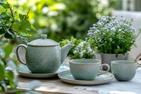 Mint green ceramic teapot and cups with textured pattern on wooden table outdoors, surrounded by blooming white flowers and fresh greenery in garden setting.