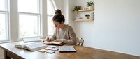 Young woman working from home at wooden desk near window with natural light, studying or writing in notebook with books and supplies for productivity.