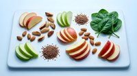 Fresh apple slices with almonds, flax seeds and spinach leaves arranged on white cutting board for healthy eating and nutrition concept.