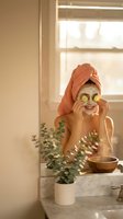 Young woman applying cucumber eye mask in bright bathroom with towel wrapped hair enjoying morning skincare routine for wellness and self-care.