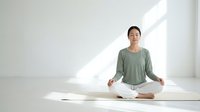 Young Asian woman practicing meditation in lotus pose on yoga mat in bright minimalist room with natural sunlight for wellness and mindfulness concept.