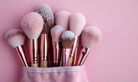 Professional makeup brushes with soft pink and gray bristles in rose gold metallic handles arranged in pink textile holder against pastel pink background.