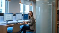 Professional businesswoman with glasses working at dual monitor computer setup in modern office environment with natural lighting and contemporary design.