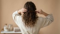 Woman with long curly hair applying treatment in bathroom, rear view showing natural hair care routine and self-care wellness lifestyle.