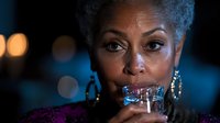 Elegant mature African American woman with silver hair sipping cocktail in evening setting, wearing purple dress and gold hoop earrings against dark background with bokeh lights.