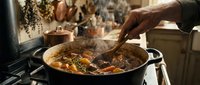 Hands stirring hearty beef stew with vegetables in cast iron pot on stove. Steam rising from homemade comfort food cooking in rustic kitchen setting.