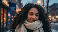 Young African American woman with curly hair wearing cozy knit scarf smiling warmly on urban street with bokeh lights during evening golden hour.