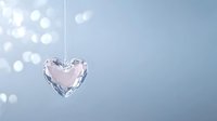 Crystal heart pendant catching light against soft blue background with bokeh effect creates romantic atmosphere for wedding and valentine designs.