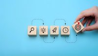 Hand placing wooden blocks with business icons showing search, innovation, growth and time management concepts on blue background for strategic planning.