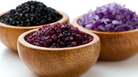 Three wooden bowls filled with colorful sea salt crystals including black, purple and red varieties for gourmet cooking and spa treatments.