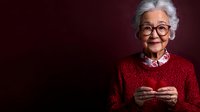 Senior Asian woman in red sweater and glasses smiling warmly while holding smartphone against burgundy background, portraying wisdom and modern technology adoption.