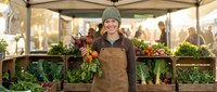 Young Caucasian female farmer in apron and beanie holding fresh carrots at outdoor farmers market stand with organic vegetables and greens display.