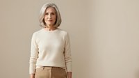 Confident mature Caucasian woman with silver bob haircut wearing cream sweater and brown pants against neutral beige background for lifestyle branding.