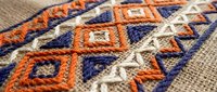 Traditional ethnic embroidery with geometric diamond patterns in orange, blue and white threads on natural burlap fabric for cultural textile design projects.