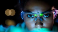 Young Asian female scientist wearing protective eyewear in laboratory setting with dramatic blue lighting and glowing reflections, close-up view focusing on determined eyes.
