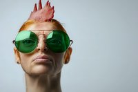 Confident young woman with pink spiked hair wearing green round sunglasses against light background, expressing individuality and modern fashion style.