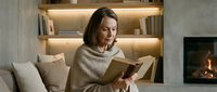 Middle-aged Caucasian woman reading book in cozy living room with fireplace and illuminated shelves, wrapped in beige blanket on sofa at home.