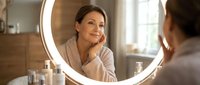 Middle-aged woman looking at herself in illuminated bathroom mirror during morning skincare routine, reflecting on self-care and wellness in modern home.