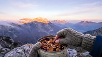 Person in gloved hands holding bowl of nuts and dried fruits on mountain peak at sunrise, alpine landscape background.