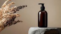 Brown glass pump bottle on concrete pedestal with dried pampas grass and natural elements against beige background. Minimalist cosmetic product presentation.